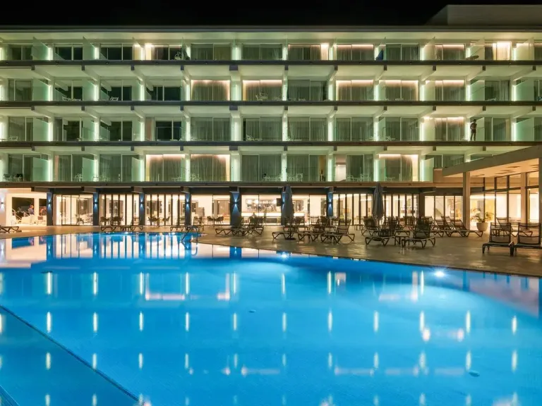 piscina grande con tumbonas frente a un hotel con balcones iluminados de las habitaciones y un gran vestibulo