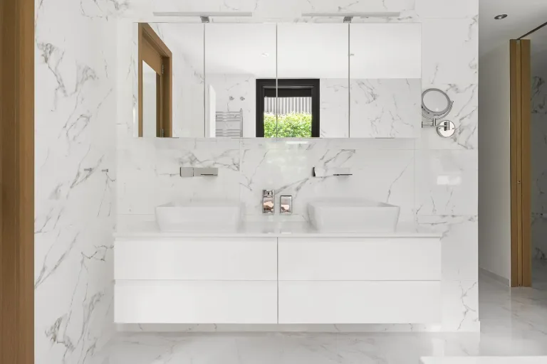 Bano pulcro con marmol blanco doble lavabo y acentos de madera. Contraste sereno y moderno