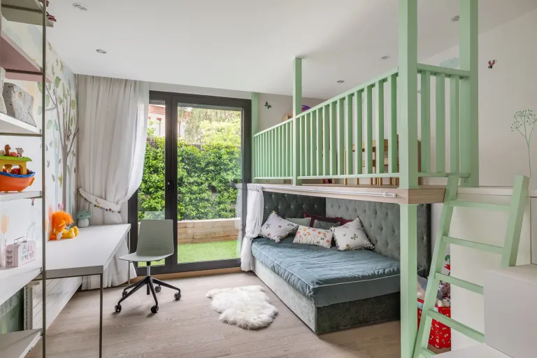 Es un espacio infantil con sofa cama gris acceso al jardin escritorio y estanterias con juguetes