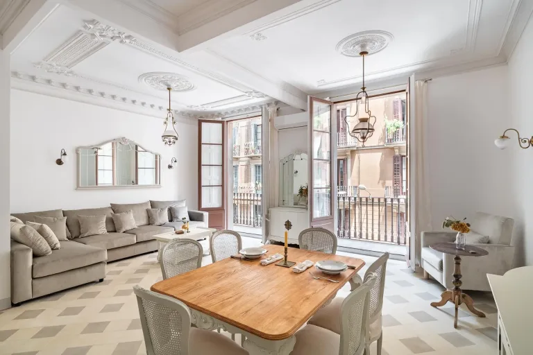 Mesa de comedor de madera en un salon con lamparas de arana un sofa sillas y dos balcones con puertas abiertas