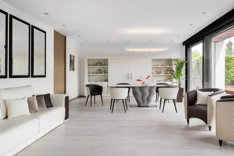 Sala de estar luminosa con sofa blanco mesa de comedor y sillas modernas junto a una vitrina y ventanal al jardin