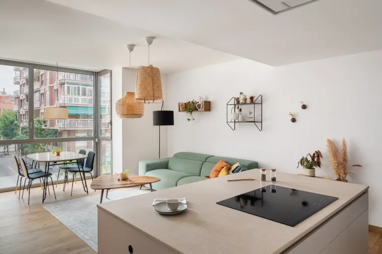 Un luminoso salon con sofa verde lamparas de mimbre colgantes y una gran ventana con vistas a edificios