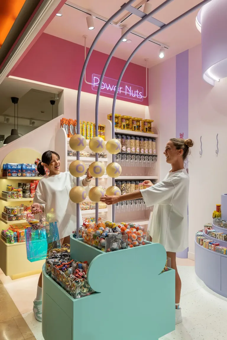 Una mujer elige dulces en una tienda con una canasta y una bolsa iridiscente junto a ella min