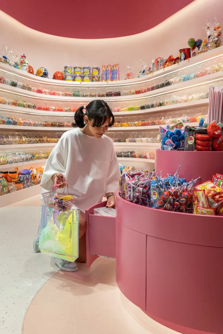 Una mujer elige dulces en una tienda con una canasta y una bolsa iridiscente junto a ella. min
