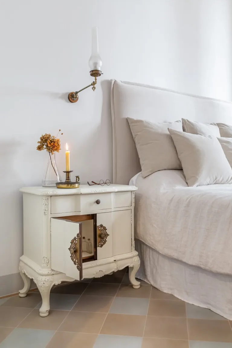 cama blanca con muchas almohadas y una mesita de noche antigua con velas y anteojos