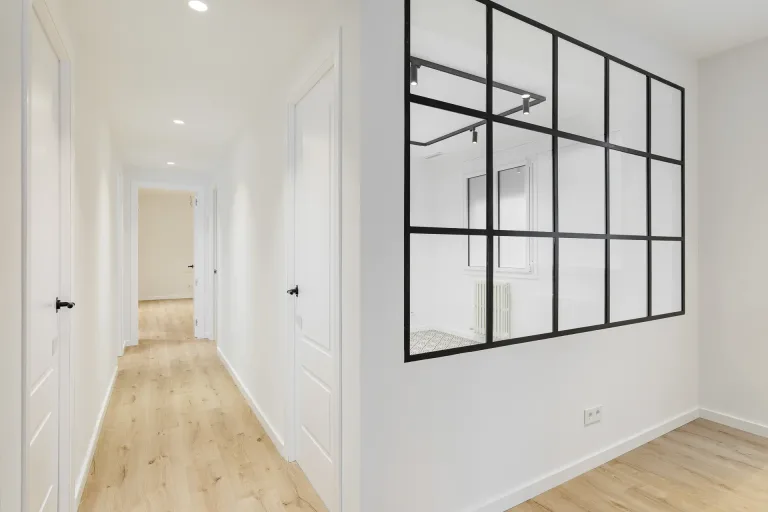 pasillo blanco con piso de madera y ventana enmarcada en negro que muestra una habitacion vacia en un apartamento nuevo