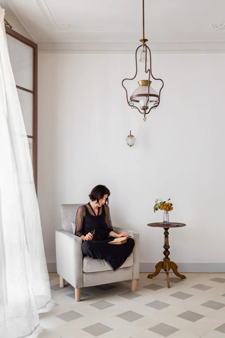 una mujer vestida de negro sentada en una silla y leyendo un libro en una sala de estar con cortina soplada