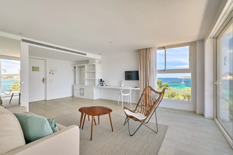 una sala de estar de un hotel con vista a mar un sofa una silla una mesa y un televisor una pared interior blanca