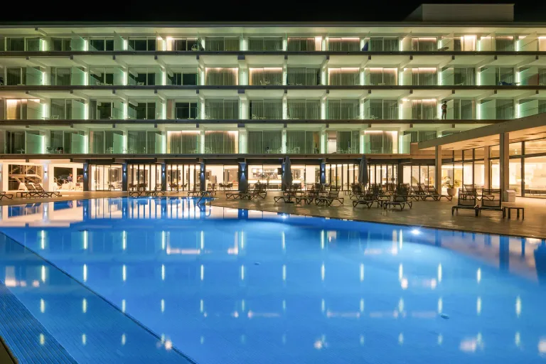 vista nocturna de una gran piscina frente a un hotel con tumbonas y mesas