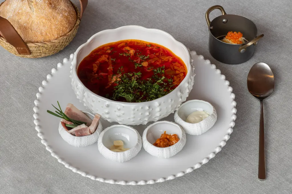 Borsch tradicional servido en un cuenco blanco con relieve acompañado de condimentos y pan de centeno