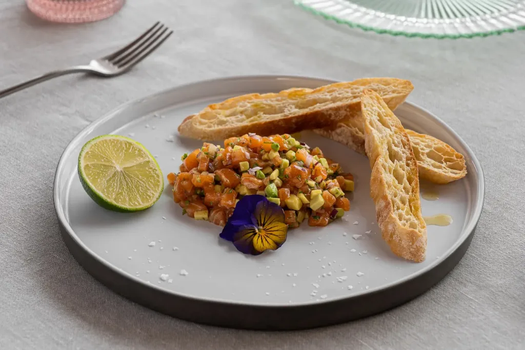 Tartar de salmón fresco con aguacate en plato gris redondo adornado con rodaja de lima y tostadas