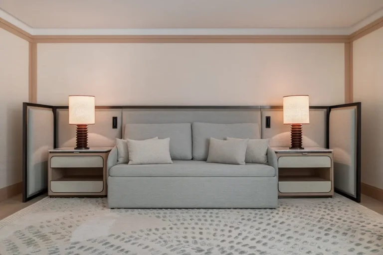 sofa gris integrado en cabecero con mesitas y lamparas en habitacion de hotel